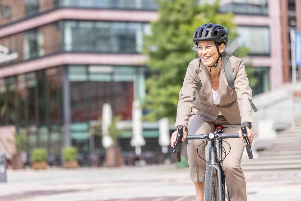 Lachende Frau mit Brille und Helm sitzt auf einem Bikeleasing Fahrrad und fährt durch die Stadt.